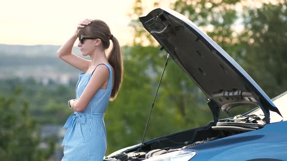 Young woman driver standing near a broken car with popped up hood waiting for help to arrive.