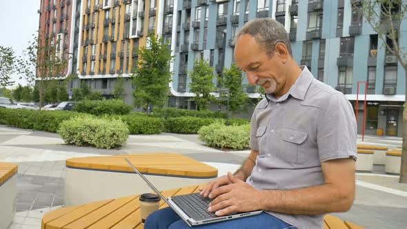 Trendy Mature Man is Working Outside with Laptop Sitting at the Bench Near New Modern Residential