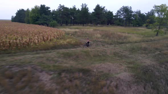 Young Sportive Woman Rides Bicycle on Countryside Road Near Corn Field and Rocks at Summer Evening