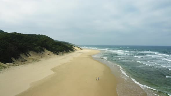 Aerial, family walks on huge sandy beach with ocean waves, cloudy sky