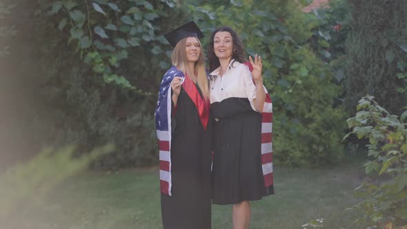 Wide Shot Cheerful Mother and Daughter in Graduation Gown Posing with American Flag Waving in Slow