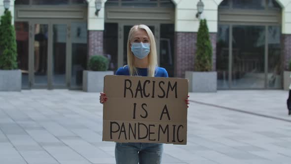 Young Woman Blonde in Medical Mask Stands with a Cardboard Poster RACISM IS A PANDEMIC in a Public