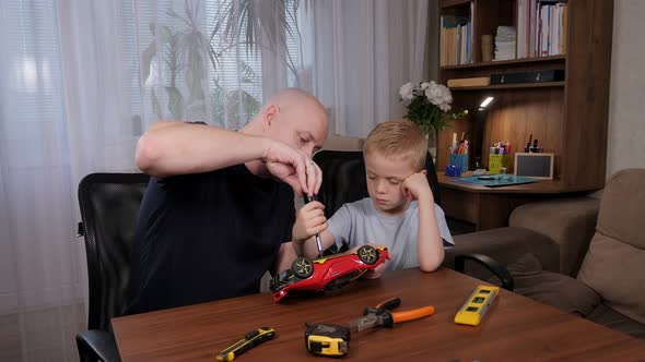 A Man and a Little Boy are Sitting at a Table and Repairing a Broken Toy Car