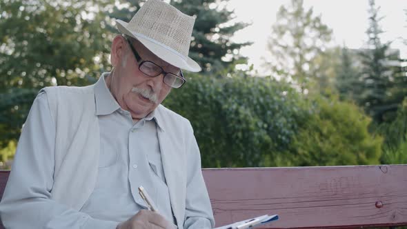 Senior Man with Glasses Sitting on Park Bench Writing on Paper and Thinking