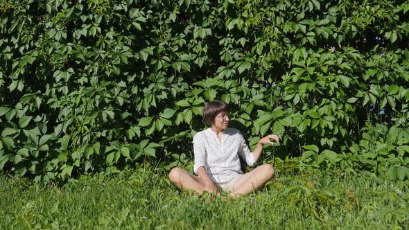 Wide Smiling Woman Sits on Grass Lawn Near Wall All Over Covered with Thick Ivy