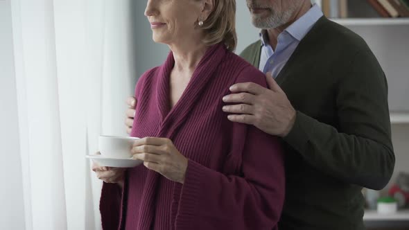 Elderly Female with Cup of Tea by Window, Man Hugging Behind, Kissing on Cheek