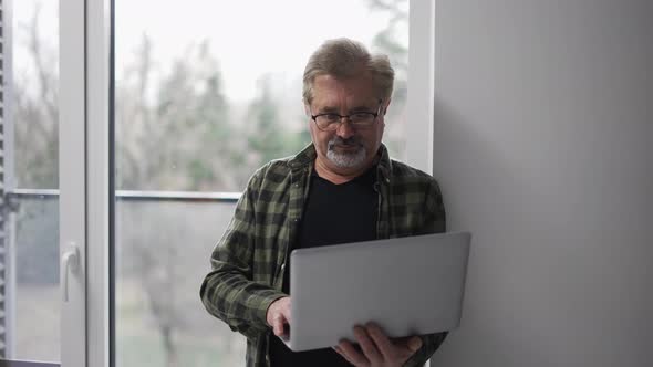 Senior Older Mature Man Typing on Laptop Browsing Internet Standing Next to the Window