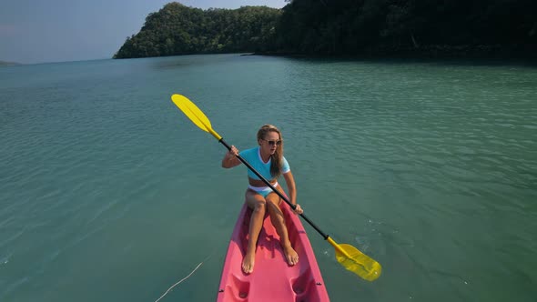 Pretty Sportive Blonde Lady in Stylish Swimsuit Sails on Pink Plastic Canoe Along Azure Ocean Bay at