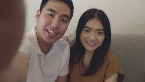 POV of young Asian couple having video chat on smartphone while sitting on the sofa at home.