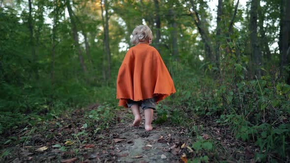 Little Toddler Boy Cosplay Gnome or Hobbit in Long Cape Walking Barefoot in Green Forest. Halloween