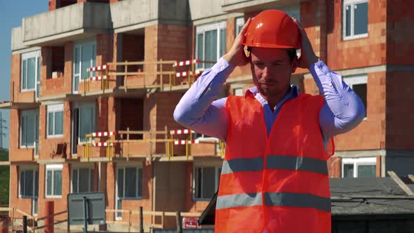 Construction Worker Puts the Helmet on His Head and Smiles To the Camera in Front of Building Site