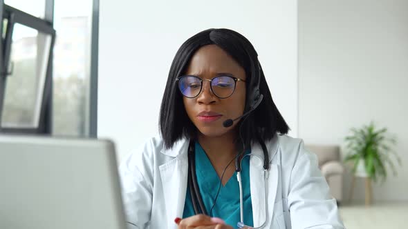 Professional African American Female Doctor in White Medical Coat and Headset Making Conference Call