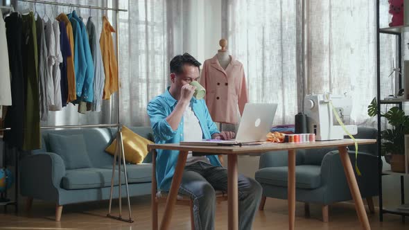 Asian Male Designer Working On A Laptop And Drinking Coffee While Designing Clothes In The Studio