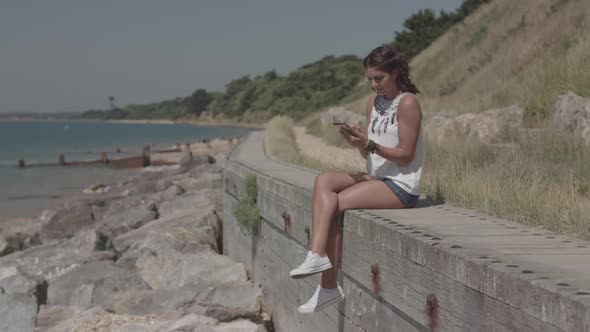 A Young Attractive Woman Sat By The Sea Using Her Phone To Order A Product Online - Ungraded