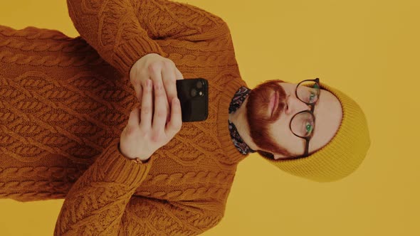 Caucasian Hipster Wearing Glasses and Beanie Using His Smartphone and Looking Around Over Yellow