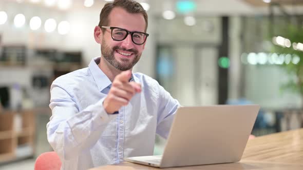 Businessman with Laptop Pointing at the Camera in Office 