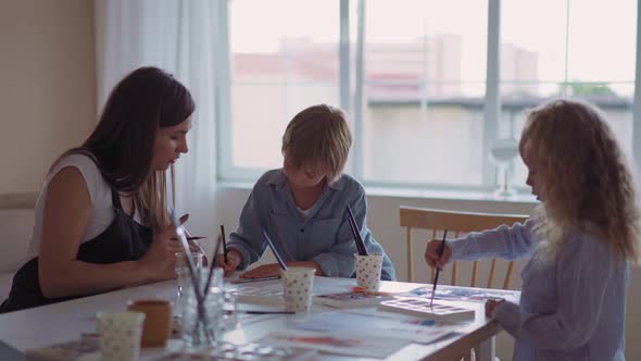 Group of Kids and Their Teacher Leaning Over Table with Watercolor Paints and Painting Selfmade Clay