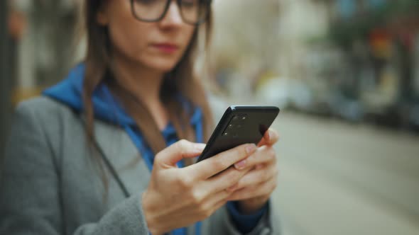 Woman with Glasses Stands at a Public Transport Stop and Uses Smartphone