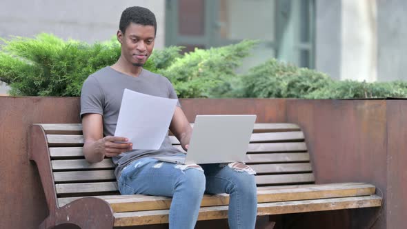 Young African Man Working on Papers and Laptop on Bench 