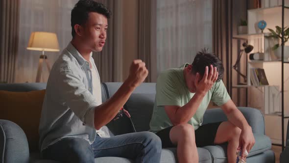 Asian Friends Playing Video Games At Home. Male Victory And Another Man Feeling Disappointed