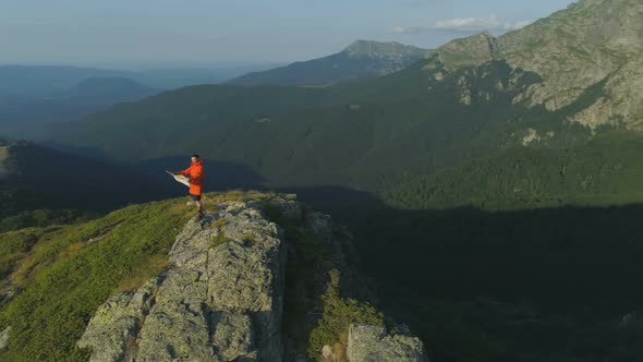 Hiker with Orange Jacket Standing on Top of the Mountain and Holding a Map Looking Around