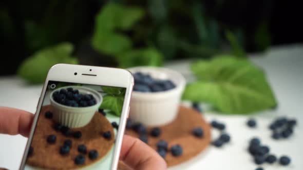 Girl Take Photo of Tasty Blueberry in White Bowl on White Table with Green Plant on Background