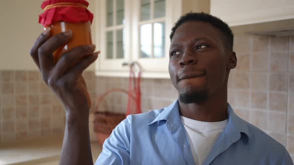 Smiling Young Man Admiring Orange Jam in Jar Standing in Kitchen Indoors