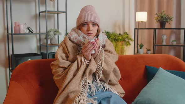 Young Sick Woman Wear Hat Wrapped in Plaid Sit Alone Shivering From Cold on Sofa Drinking Hot Tea