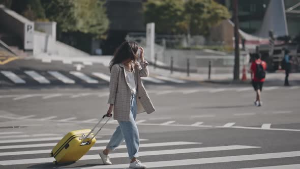 Portrait of Young Woman Walkinig Across the Street Towards Camera Rolling Suitcase Behind