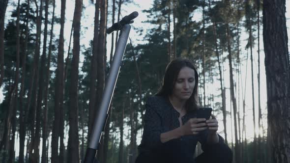 Young businesswoman in suit using smartphone while sitting on electric scooter in park at sunset.