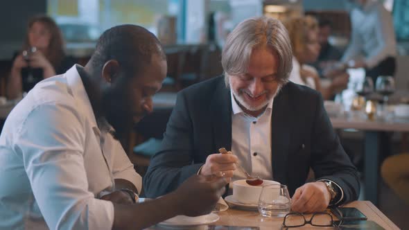 Smiling Mature Businessman Sitting at Cafe with African Partner Enjoying Meal.