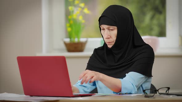 Concentrated Mature Woman in Black Hijab Typing on Laptop Keyboard Sitting at Table Indoors