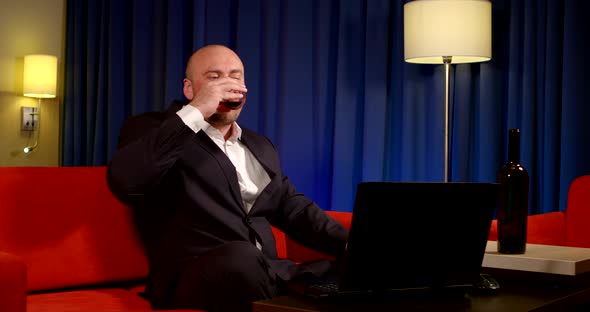 a Bald Man in a Dark Suit Sits on a Red Sofa Against a Blue Curtain. He Talks Cheerfully Over a