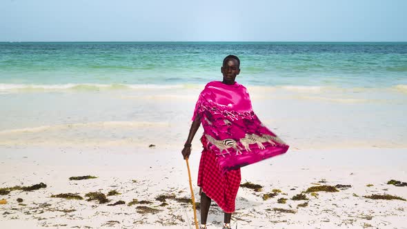 Native african man in pink clothing holding stick stands on sand beach.