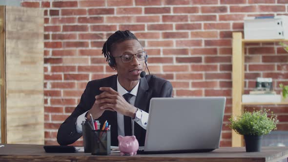 Wearing a Headset and Glasses a Black Man in Suit Speaks Via Video Link Through Laptop at an Office