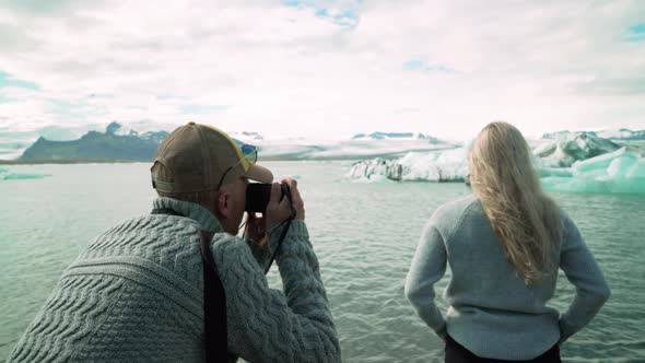 Adult Man Taking Pictures Of Woman Posing With Icebergs In The Background At Jokulsarlon Glacier In