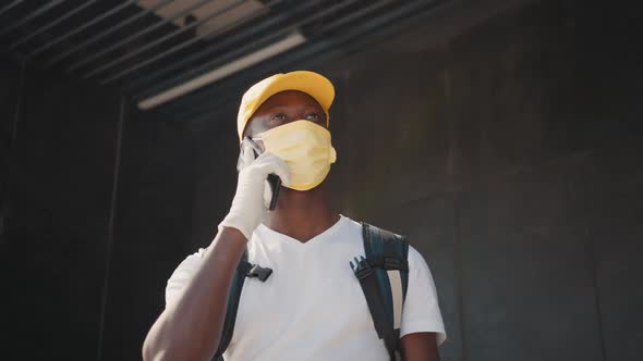 A Young Black Courier in a Mask and Gloves Talks on the Phone with a Customer