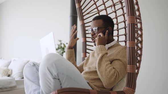 Angry AfricanAmerican Man Shouts Into the Phone While Sitting in a Suspended Chair with a Laptop on