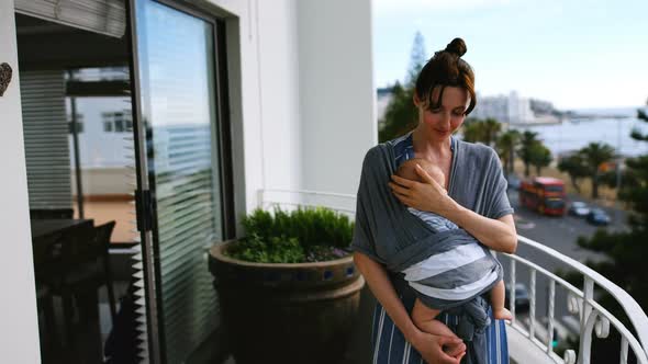 Young smiling mother standing in balcony with baby in sling 4k