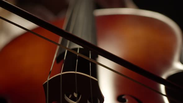 Bow plays slowly on cello strings, against a dark background. Light touches of bow to strings