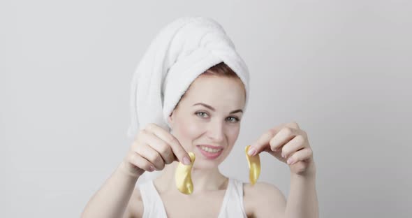 Smiling Woman with Hair Wrapped in Towel Posing with Golden Under Eye Patches in Hands