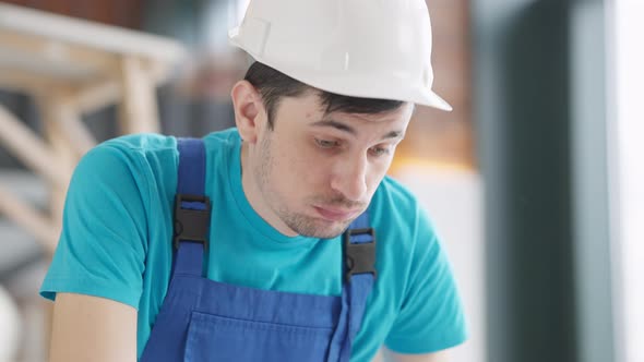 Closeup Portrait of Thoughtful Tired Young Service Man in Hard Hat Wiping Forehead with Hand Sighing