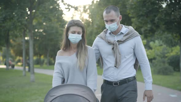 Young Caucasian Man and Woman in Covid Face Masks Strolling at Sunset with Baby Carriage and Talking