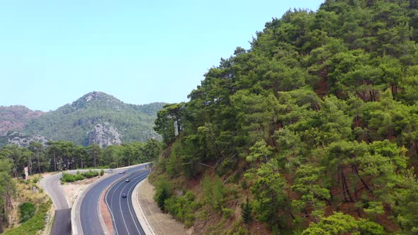 aerial drone flying over a mountain hill filled with trees next to a highway as cars pass by on a su