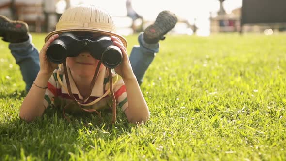 Child Wearing Pith Helmet is Playing in Summer Day with Binoculars Lying Down on the Grass at Town