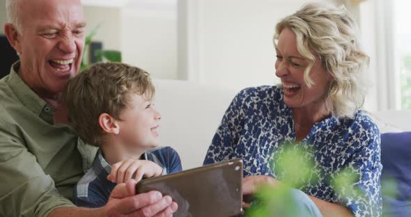 Happy caucasian grandparents with grandson using tablet and sitting in living room