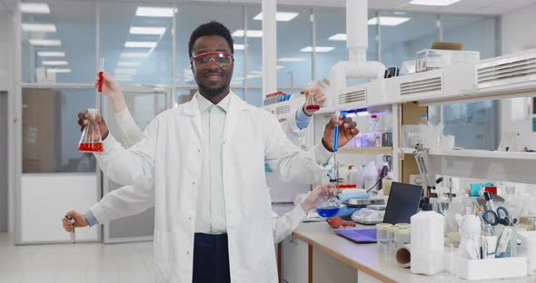 Multihanded Afro Laboratory Scientist in Lab Coat and Safety Goggles Holding Flasks and Test Tubes