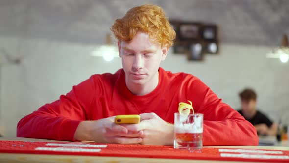 Absorbed Redhead Young Man Using Smartphone Sitting at Bar Counter with Cocktail Glass As Man