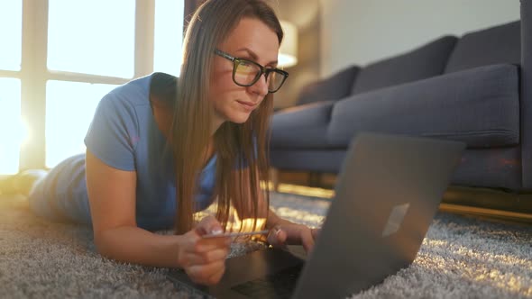 Woman with Glasses Is Lying on the Floor and Makes an Online Purchase Using a Credit Card and Laptop