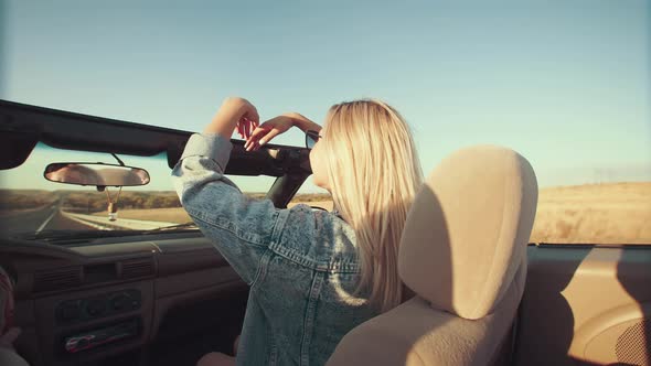 Young Woman Raises Her Hands Above Her As She Sits in Front Seat of Cabriolet